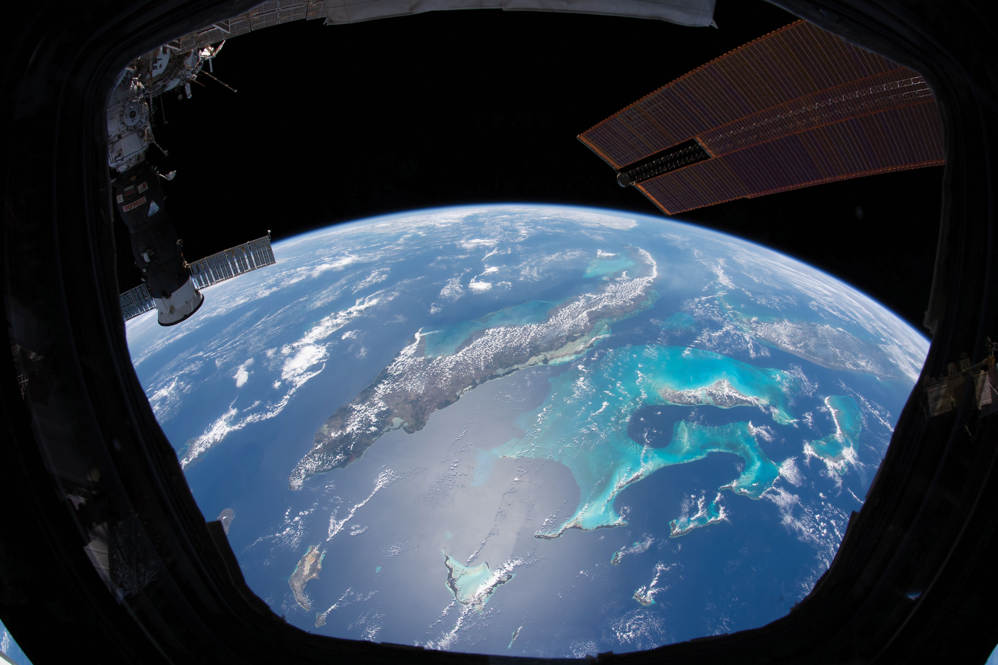A photo taken from the International Space Station shows the curving horizon of Earth below. In view is a brilliant blue ocean region with islands and small white clouds. Pieces of the station like the solar arrays are visible at the top of the photo.