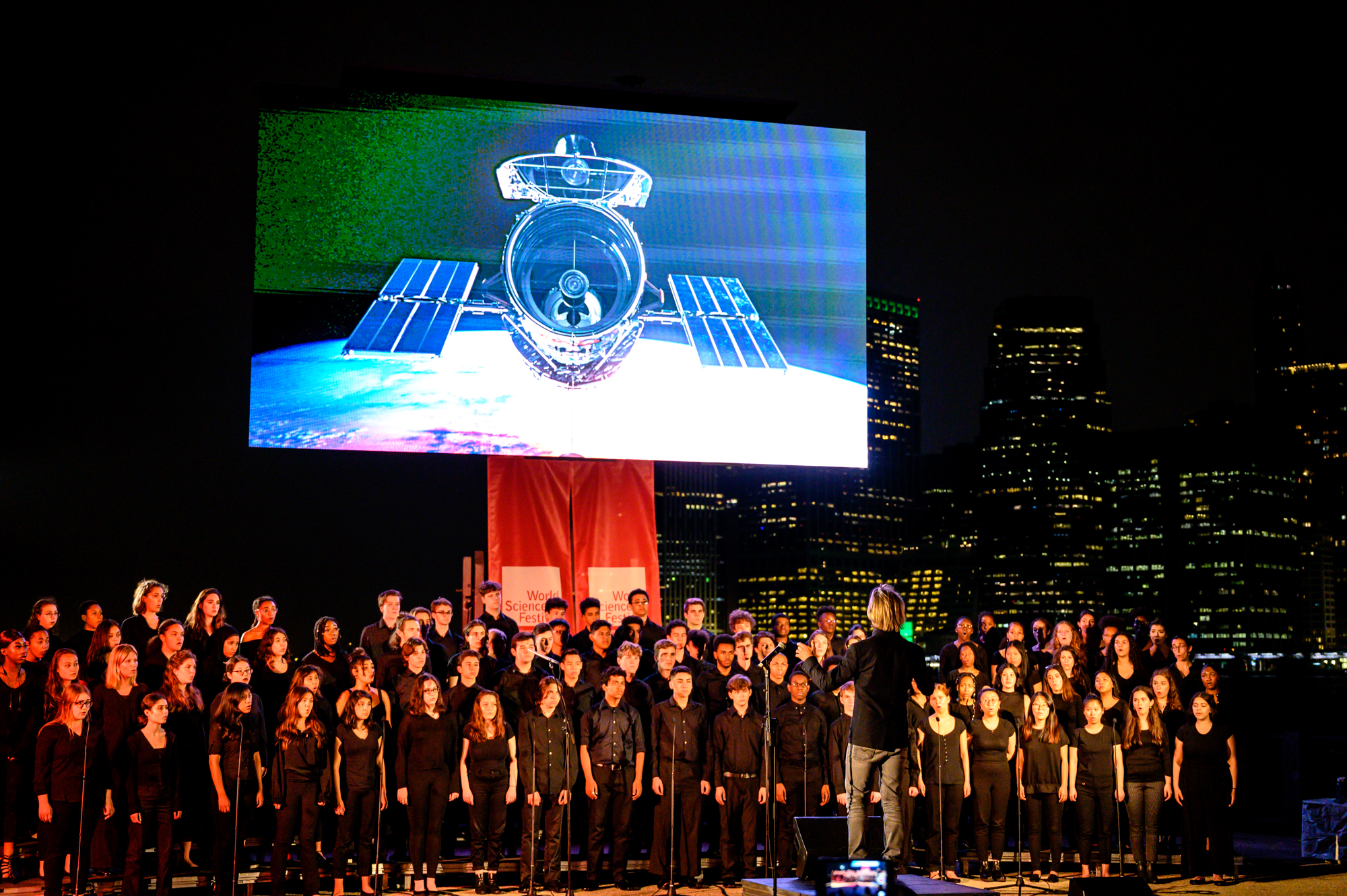 A large choir performs beneath a screen showing an image of the Hubble Space Telescope. It is night and the lit buildings of New York City are visible in the background.