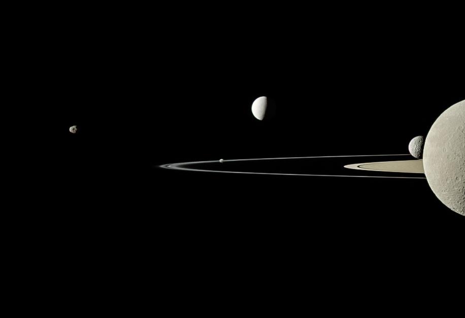 Against a black background, five golden-gray moons of varying sizes are scattered along a horizontal band through the image. Sunlight illuminates them from the left, and their right sides are in shadow. The largest, at far right, takes up the middle third of the photo, fully sunlit, with its dark half cut off by the right edge of the frame. The rings of Saturn, seen nearly edge-on, pierce the image like a knife blade from center-right, emerging through the largest moon, and with the smallest moon resting atop the blade like a breadcrumb.