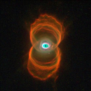 Two sets of rings in orange and gold stacked one above the other form the shape of a hourglass. At image center, where the two sets of rings overlap, is an area of white, green and black dust in a shape that is similar to the human eye.