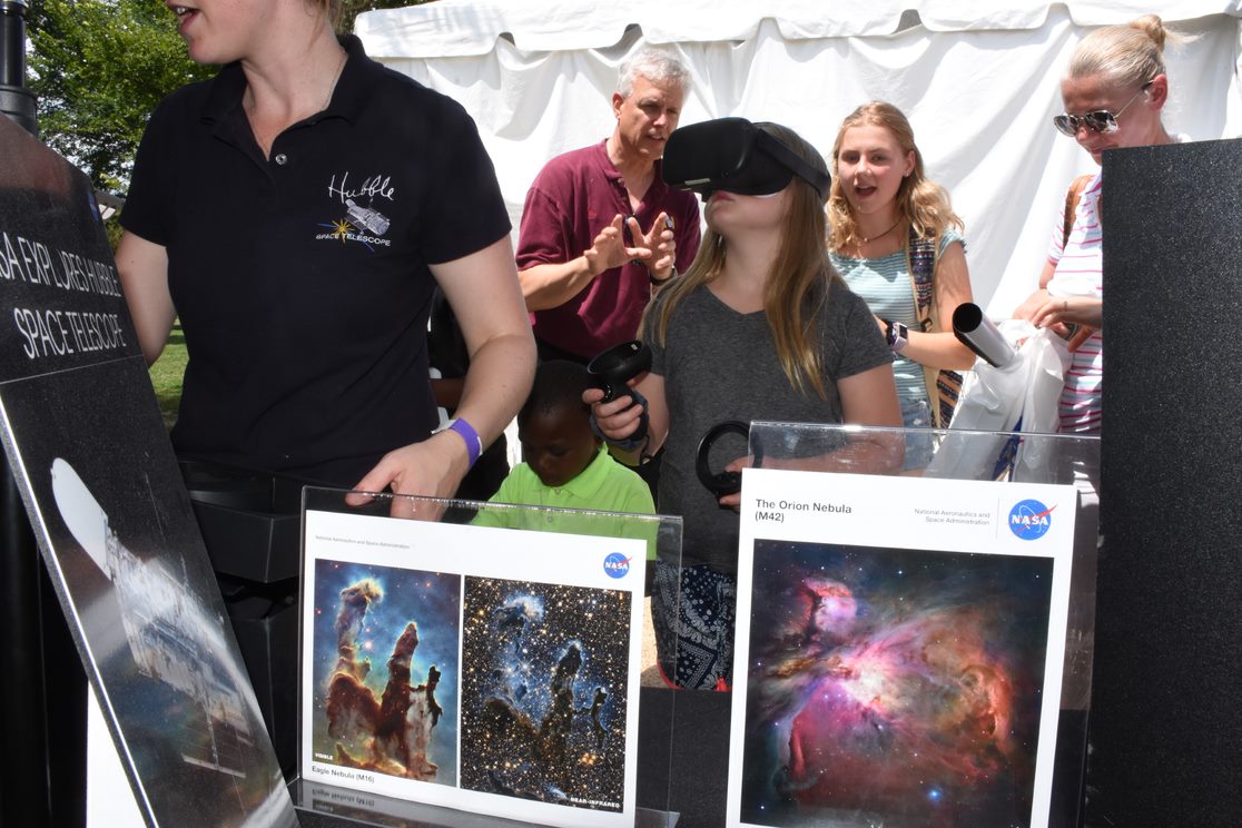 A girl wears a VR headset and holds VR controlers in her hands at a crowded outdoor event with Hubble images on a table in front of her.