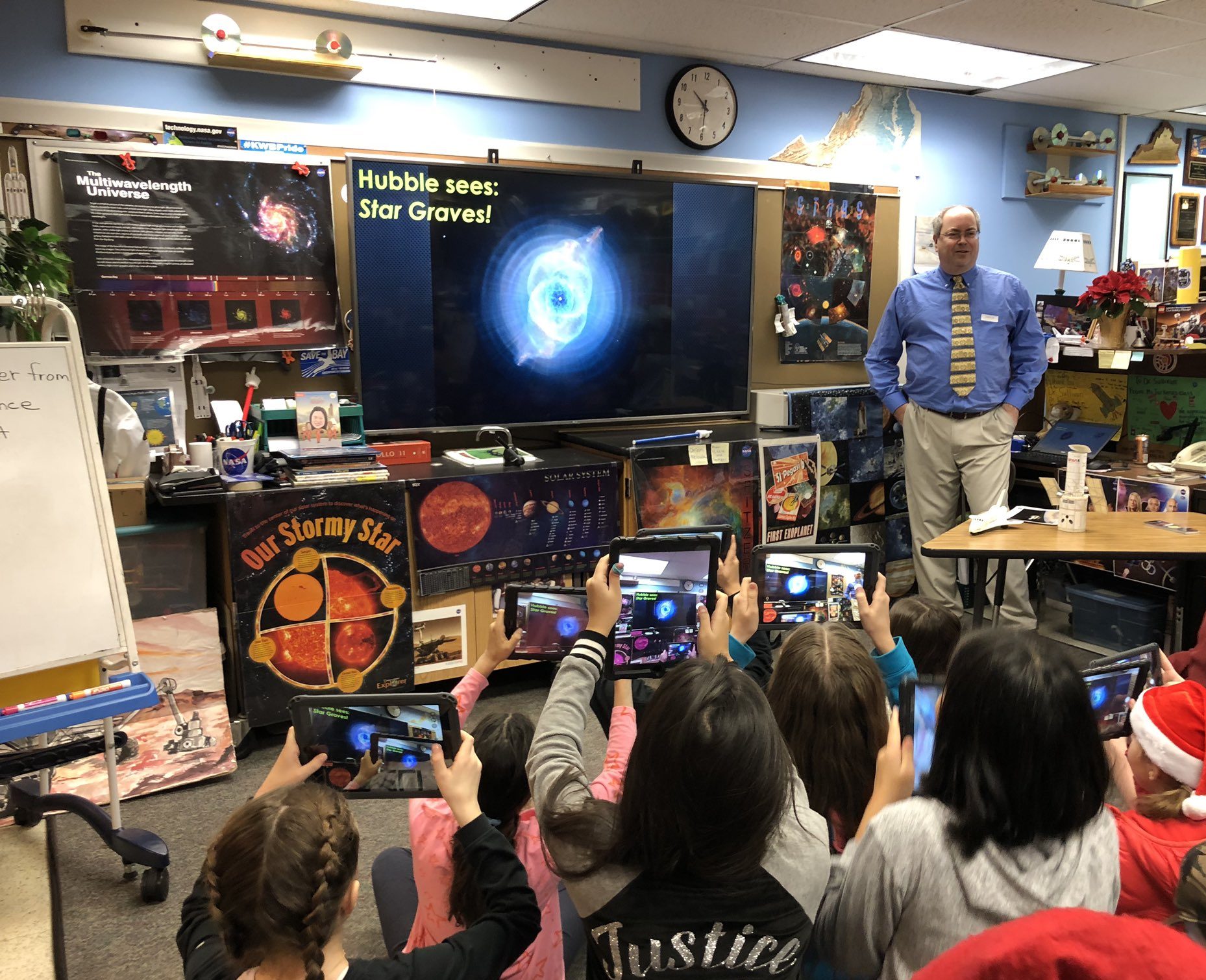 Middle-school children seated on the floor of a classroom point tablets toward a Hubble image of the Cat's Eye Nebula on a large screen in the front of the classroom. A Hubble scientist stands next to the screen discussing the image. The walls and nearby desks are covered with space and astronomy images.
