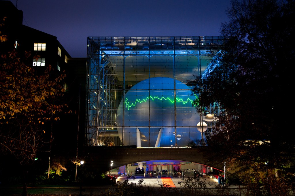 A jagged, EKG-like spectra line from the Hubble Space Telescope is projected onto a large sphere seen through a windowed wall at Hayden Planetarium