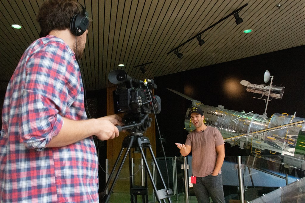 Actor Josh Peck in a t-shirt and baseball hat points into a camera operated by producer Paul Morris while delivering a birthday message. Morris is in the foreground, behind the camera, and Peck is positioned in front of a large model of the Hubble Telescope.