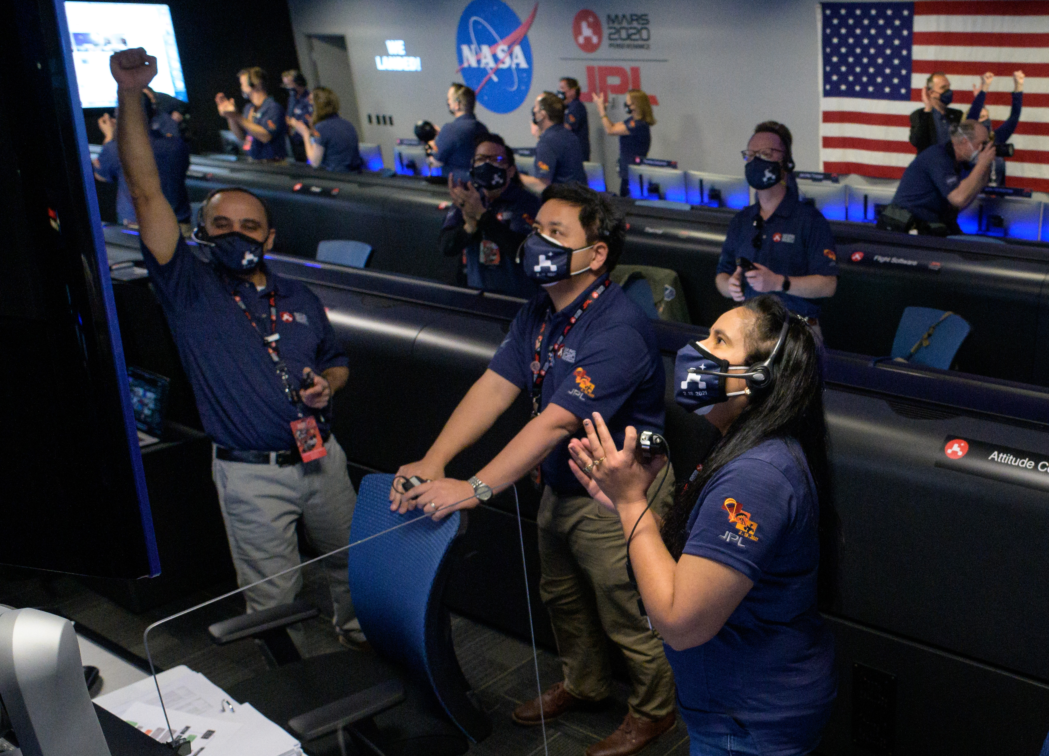 Men and women dressed in blue shirts with mission logos cheer as they watch a TV with images beamed back from Mars.