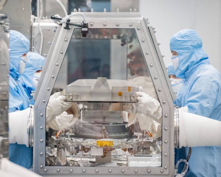 A silver capsule is opened inside a glovebox surrounded by technicians gowned in blue protective suits