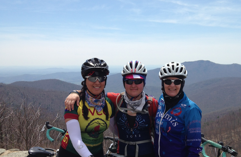 Three women in full biking gear stand together for a photo in front of a mountain range.