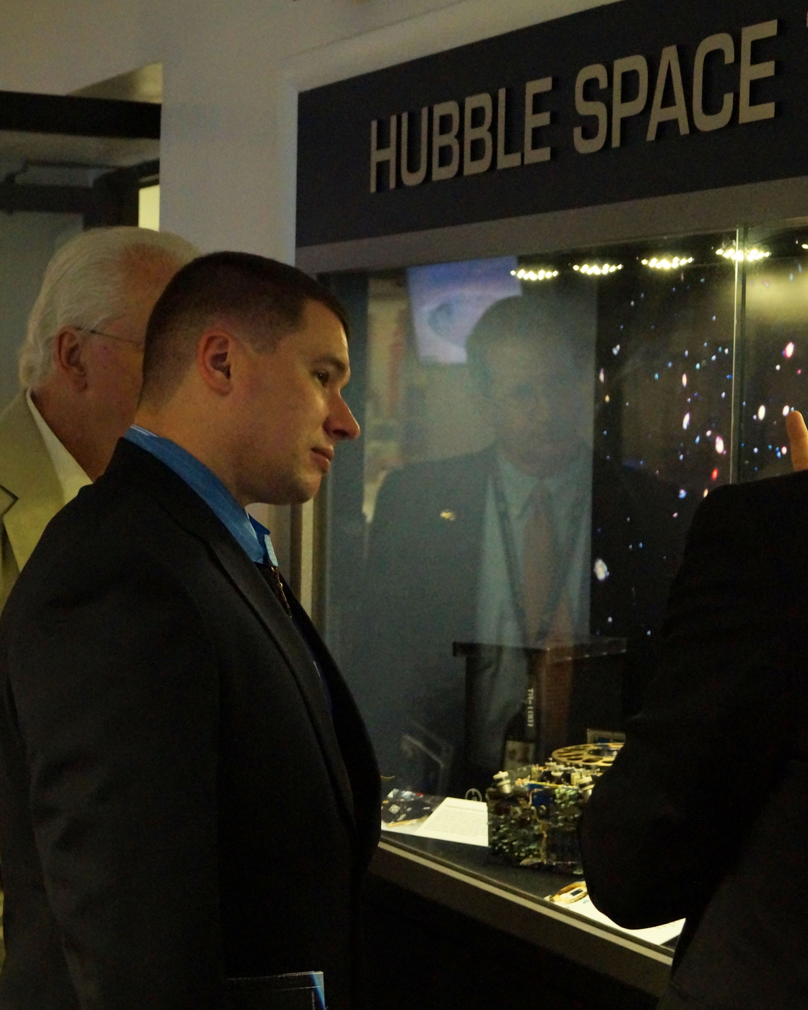 Kyle White in a dark suit gazes through a lit display case at Hubble hardware. An image of one of Hubble's deep fields, filled with stars and galaxies, is the backdrop for the case.