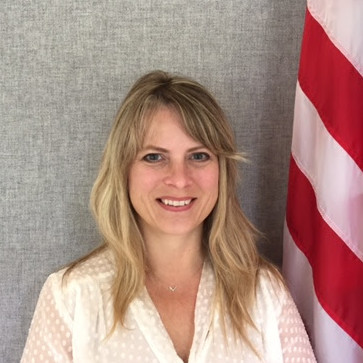 Portrait photo of a smiling woman with long blond hair standing next to the American flag.