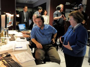Senator Barbara Mikulski gestures as she speaks with a member of the Hubble flight operations team. He sits at a long desk with another team member. The desk is covered with computers, keyboards, stacks of paper bound together with o-rings so it can be flipped through easily, and a small Hubble model. Other figures are visible in the background, including someone holding a boom mic to capture their interaction.