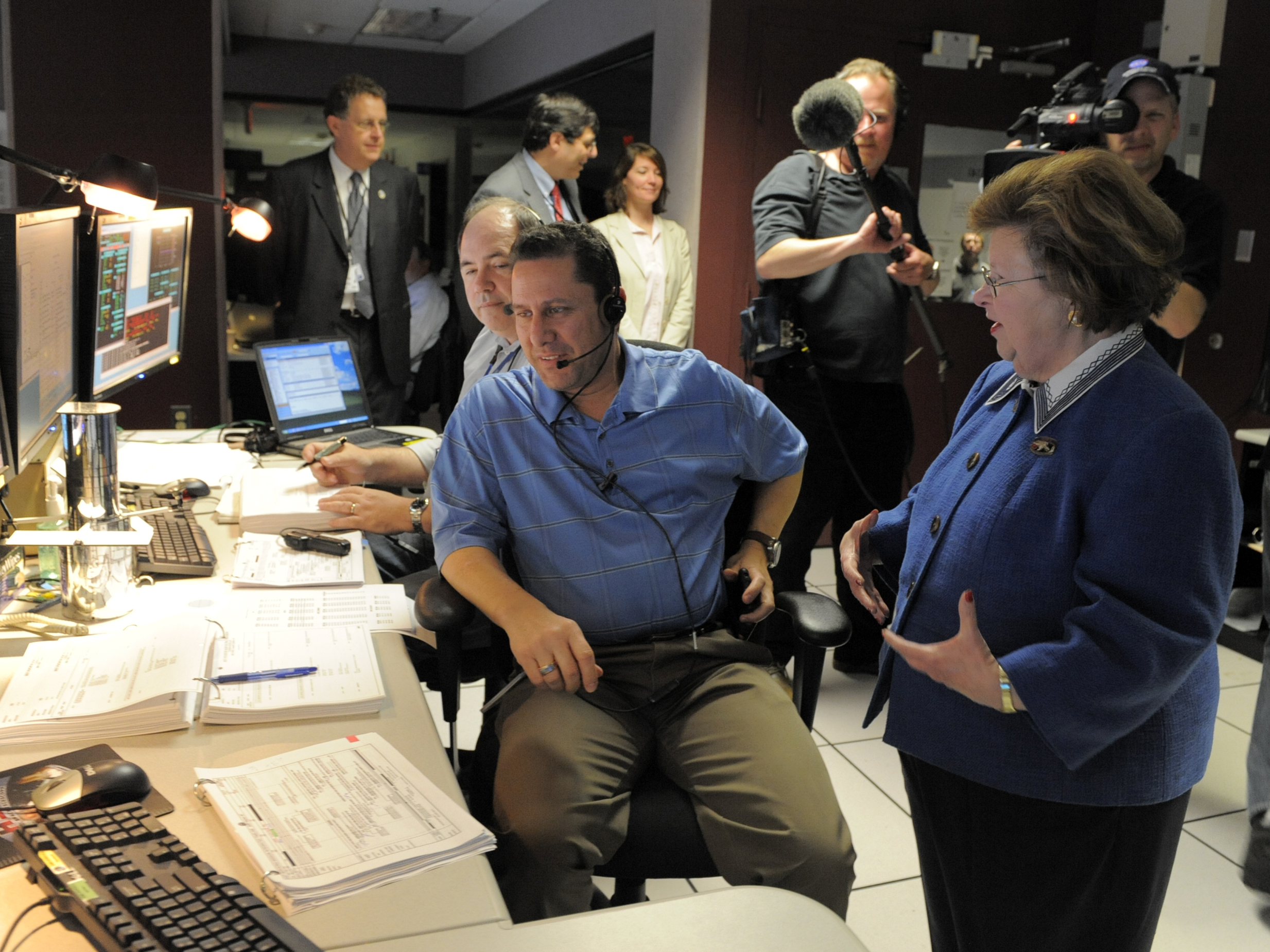 Senator Barbara Mikulski gestures as she speaks with a member of the Hubble flight operations team. He sits at a long desk with another team member. The desk is covered with computers, keyboards, stacks of paper bound together with o-rings so it can be flipped through easily, and a small Hubble model. Other figures are visible in the background, including someone holding a boom mic to capture their interaction.