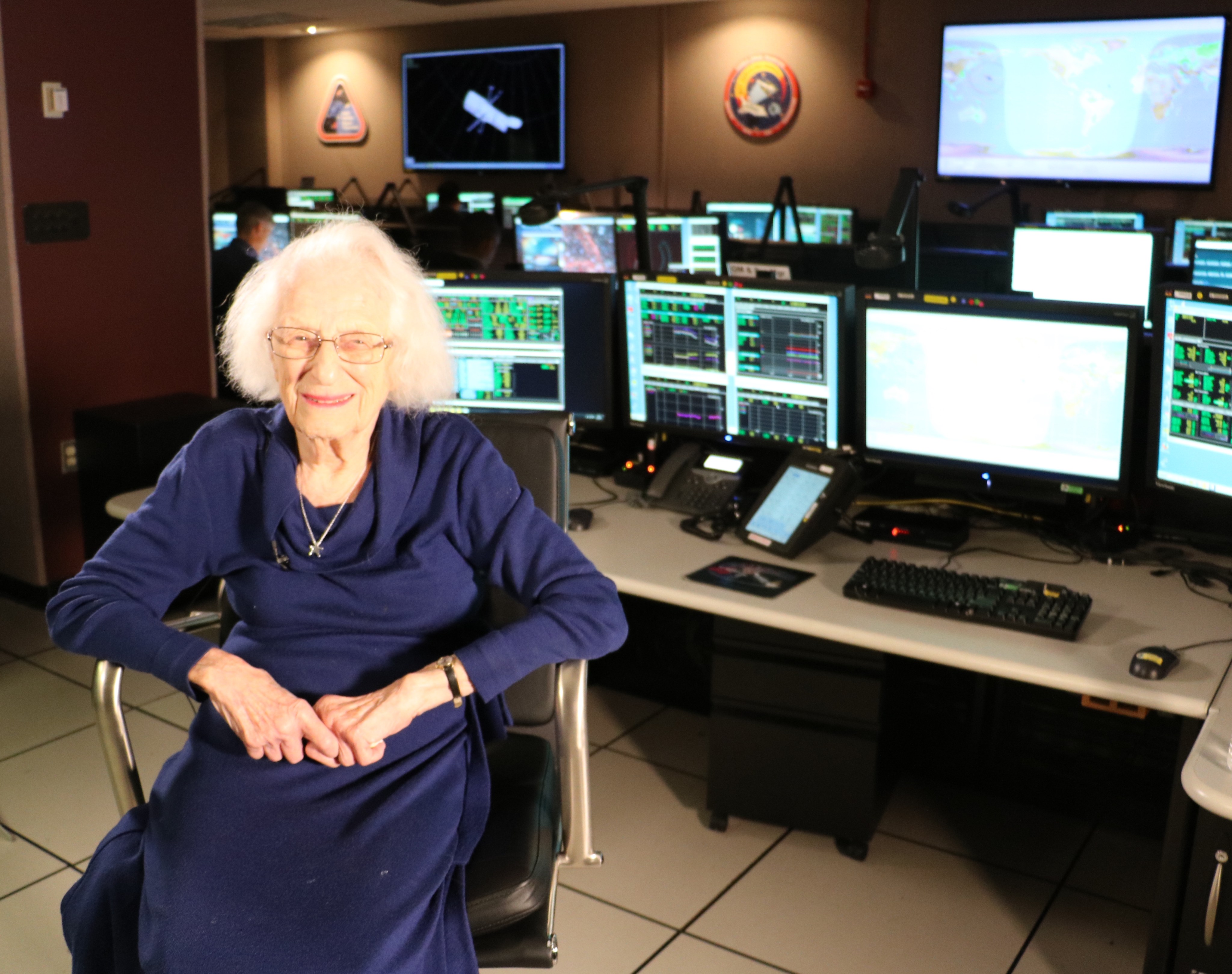 Nancy Grace Roman in a blue outfit and glasses smiles as she sits in a chair in the Hubble control room in front of desks full of computers with screens showing Hubble data.