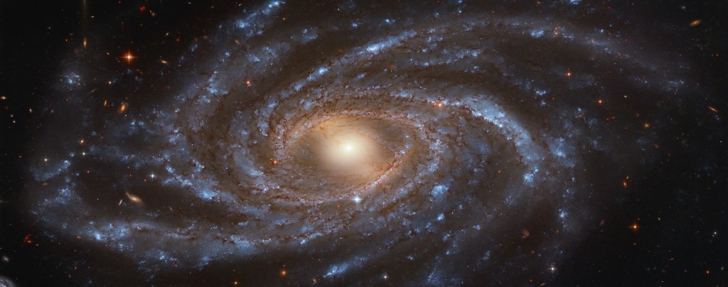 A spiral galaxy with a glowing yellow-orange center, pink swirls near the center, and light blue-white spiral arms of stars swirling out in all directions. Many more reddish galaxies can be seen faintly in the background, appearing much smaller.