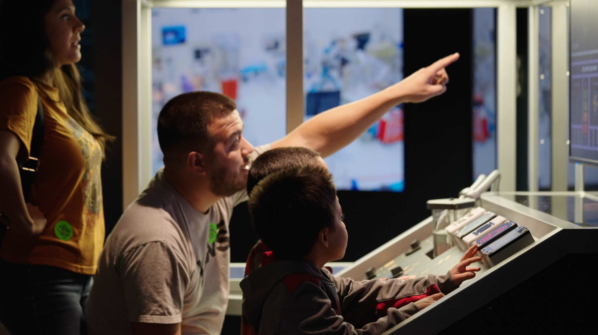 Photo of a man and child sitting at a control panel, the main is pointing at something on large screen.