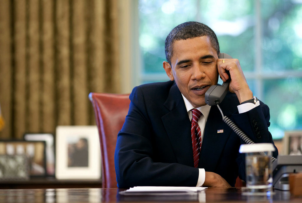 President Barack Obama speaks into a phone. He is seated at his desk in the Oval Office.