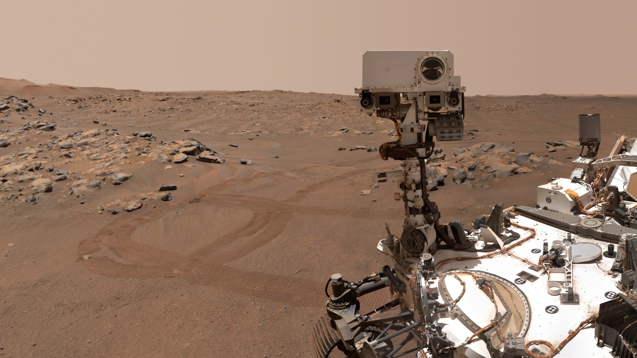 The Mars rover Perseverence is seen at right, with the rust-colored, dusty and rocky surface of Mars in the background.