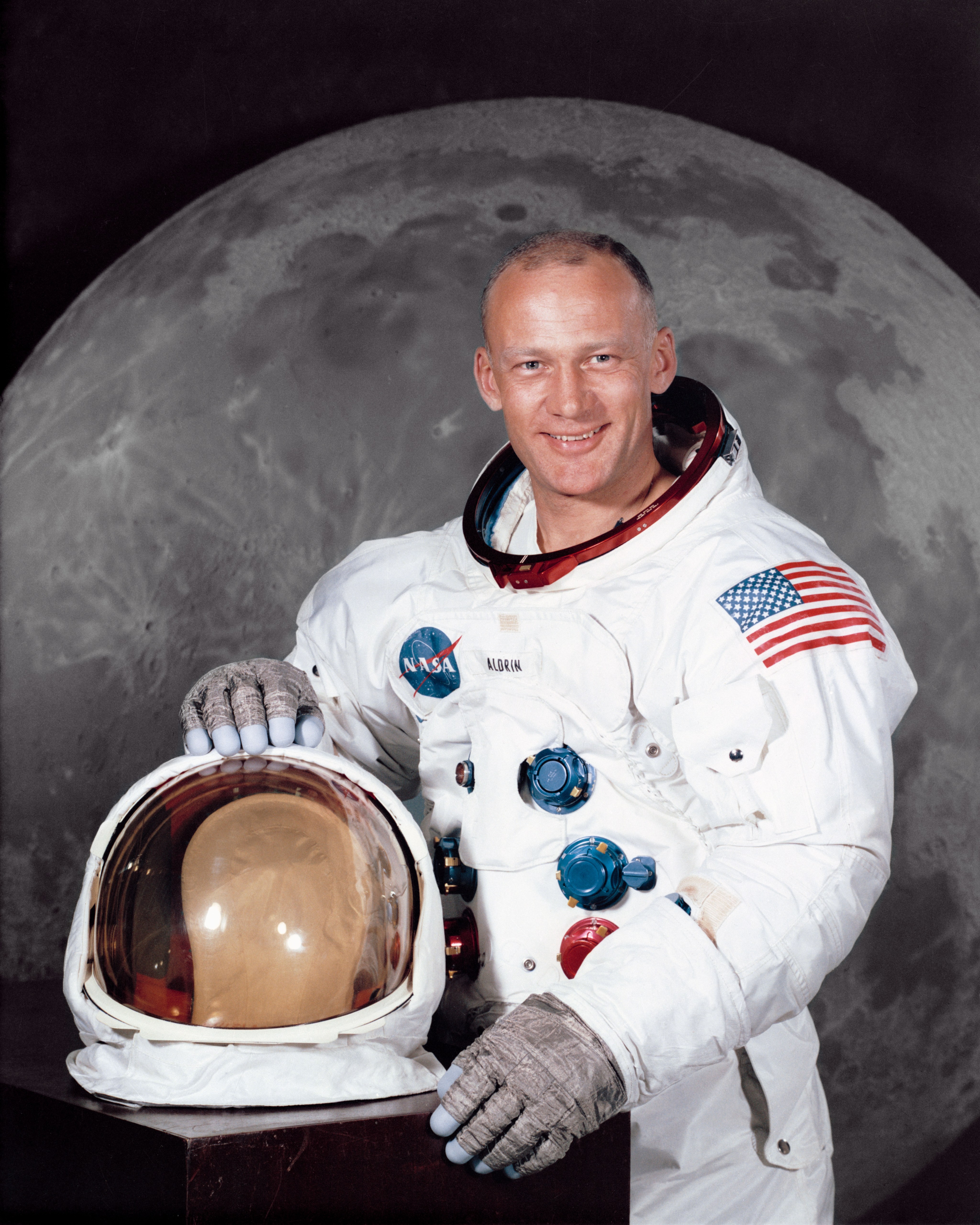 Headshot of man in a white space suit holding a helmet in front of a backdrop photo of the moon