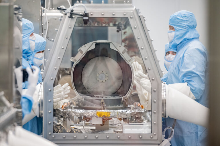 A silver capsule is opened inside a glovebox surrounded by technicians gowned in blue protective suits