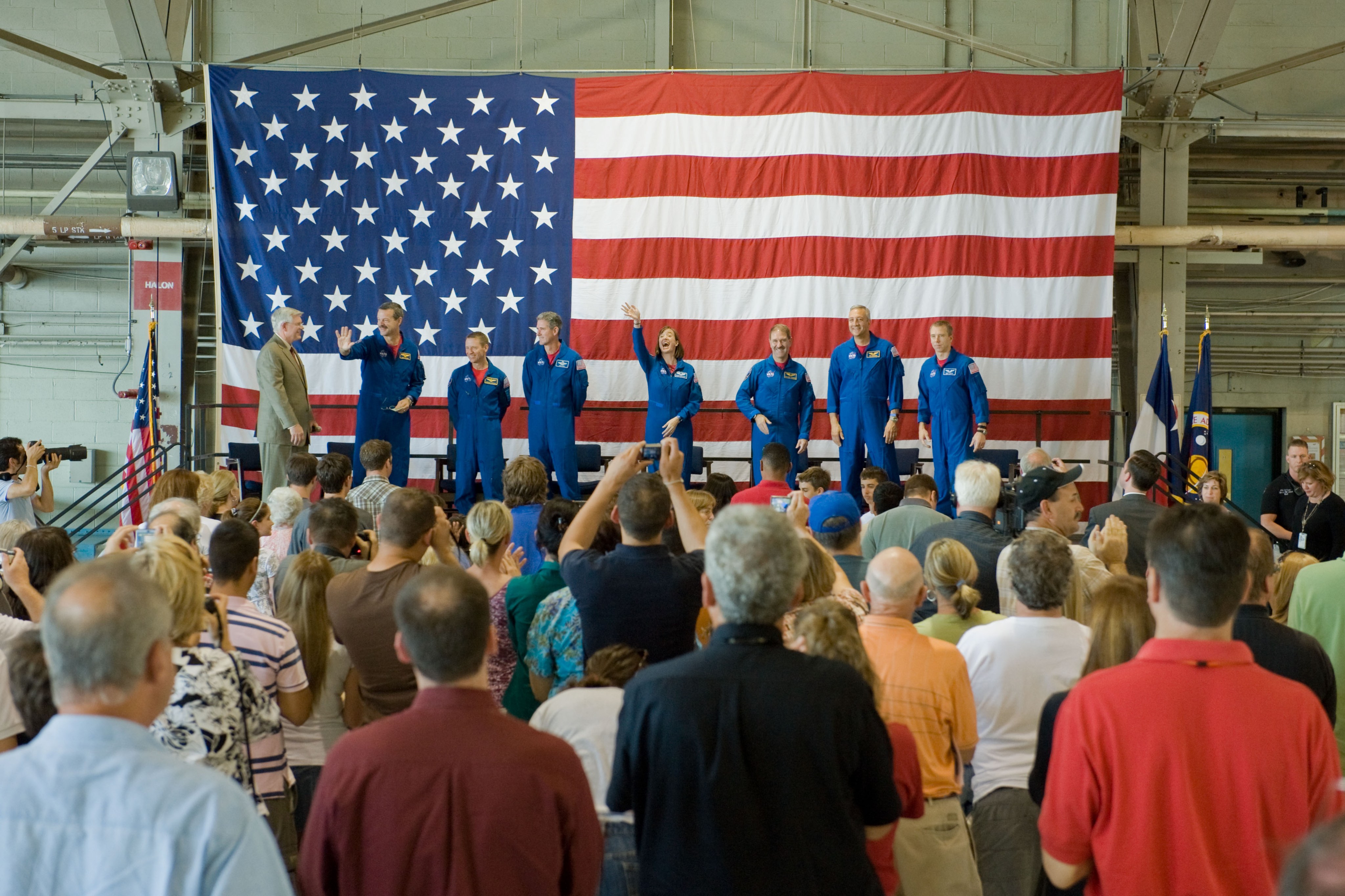 The astronauts of Servicing Mission 4 are lined up on stage with the background of an American flag, which hangs on the wall behind them. they are smiling and waving. A crowd of people are visible in front of them.