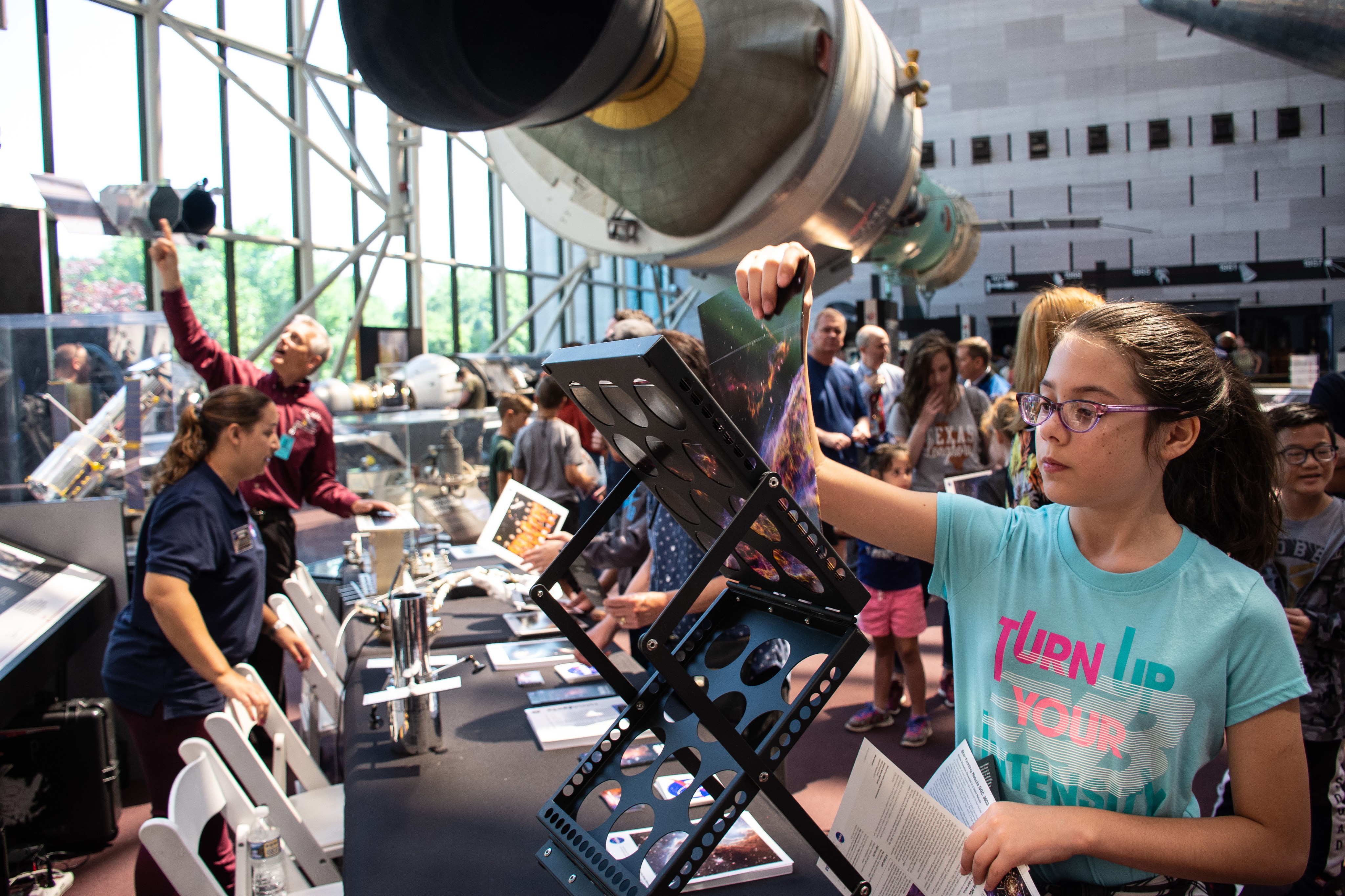 A girl selects Hubble images to take home from a table covered with Hubble activities. A large model of Hubble is visible in the background alongside other spacecraft exhibits at the National Air and Space Museum.