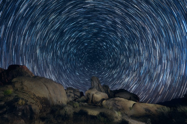 Star trails in the sky swirl above a rugged landscape.