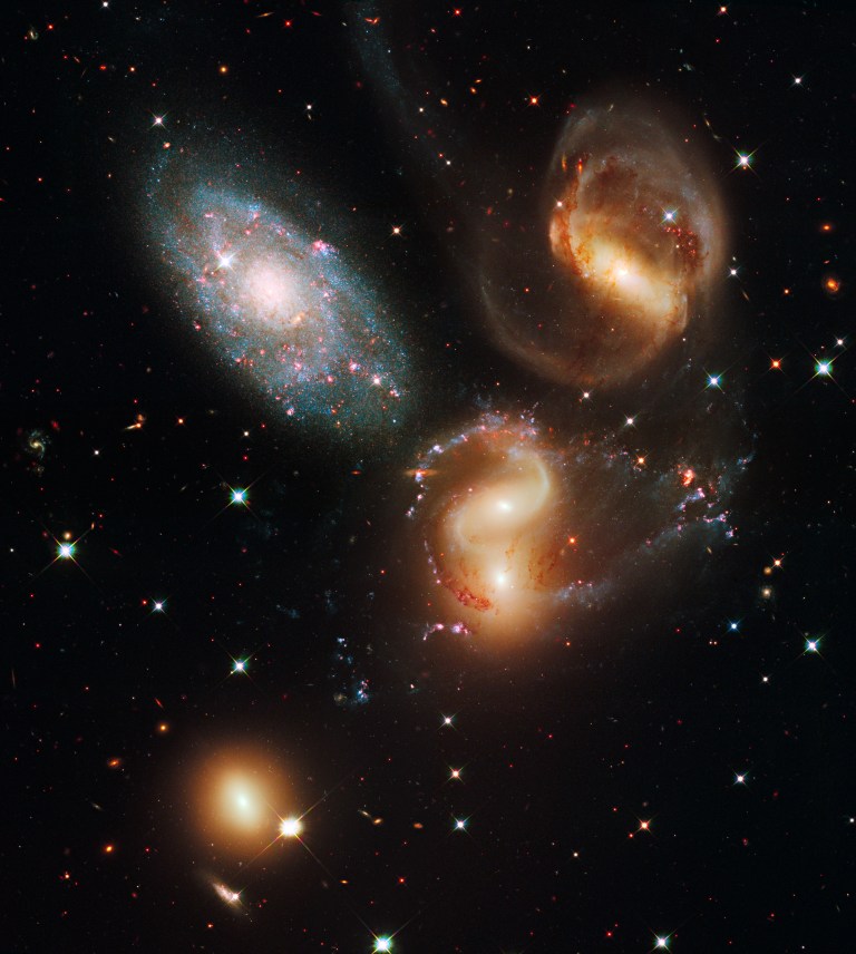 A Hubble image of 5 galaxies, three spirals in a triangle near the top (two red-orange, one blue-white), the last two (a red-orange sphere and a small distant spiral) near the bottom.