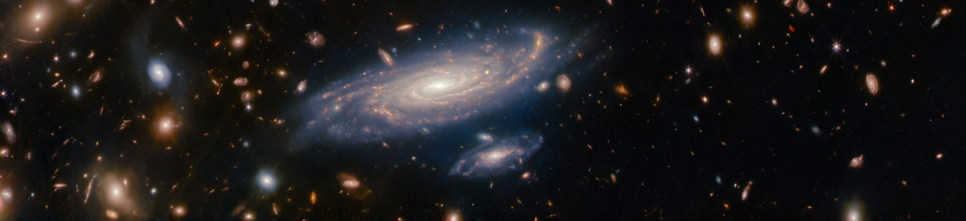 A crowded field of galaxies is interspersed with bright 8-pointed stars on a dark background. The galaxies and stars come in a variety of sizes and colors, ranging from bluish white to orange. Some galaxies are large enough to make out spiral arms, while others look like faint smudges or pinpricks. The most prominent feature is a large, detailed spiral galaxy called LEDA 2046648, seen at an oblique angle towards the bottom of the frame. A smaller spiral galaxy is just below it. About one-quarter the size of its larger companion, this small galaxy looks like a miniature version of LEDA 2046648. Both of these spiral galaxies have glowing cores and areas of star formation lighting up their pale pink arms.