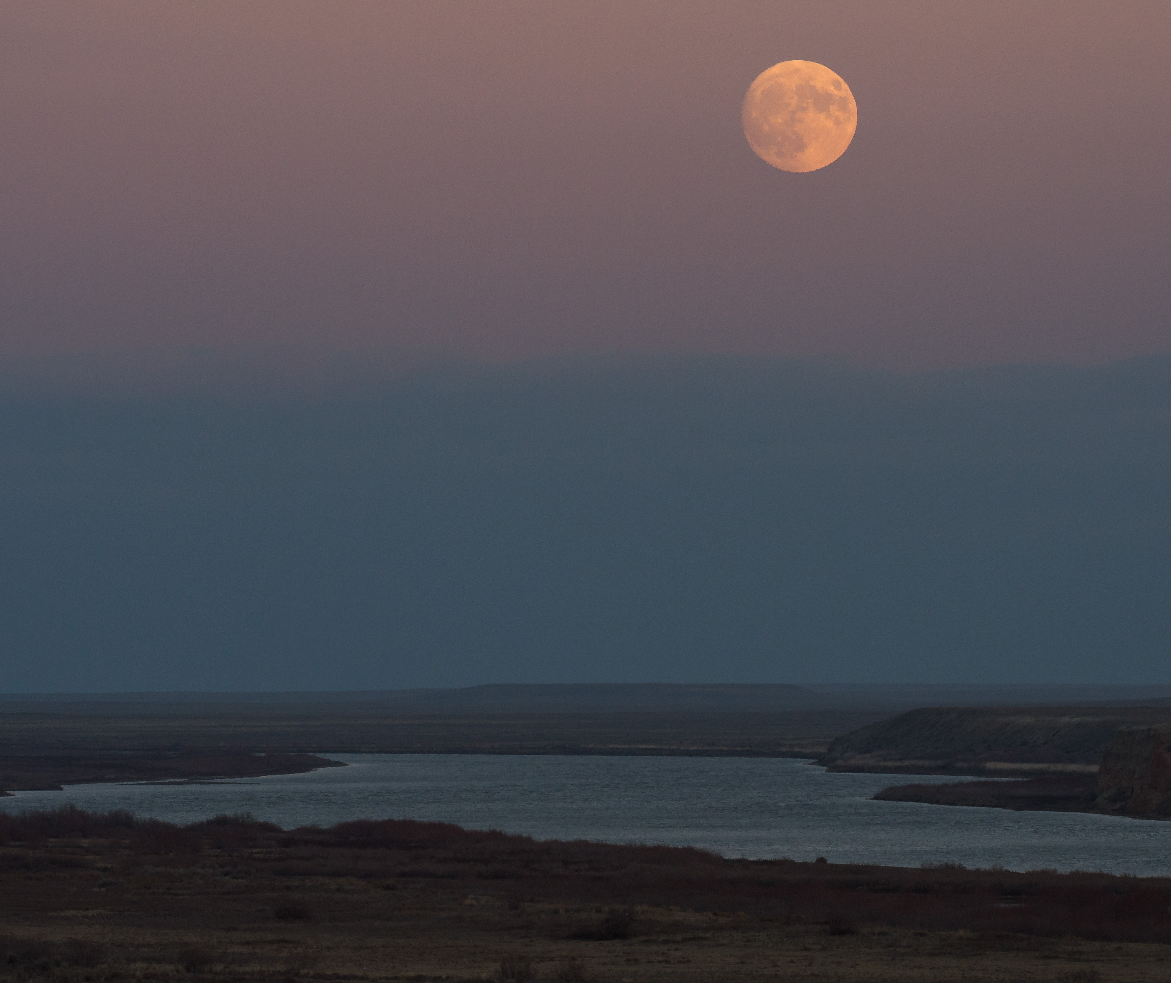 An orange full moon hangs in a pink and blue sky above a flowing river.