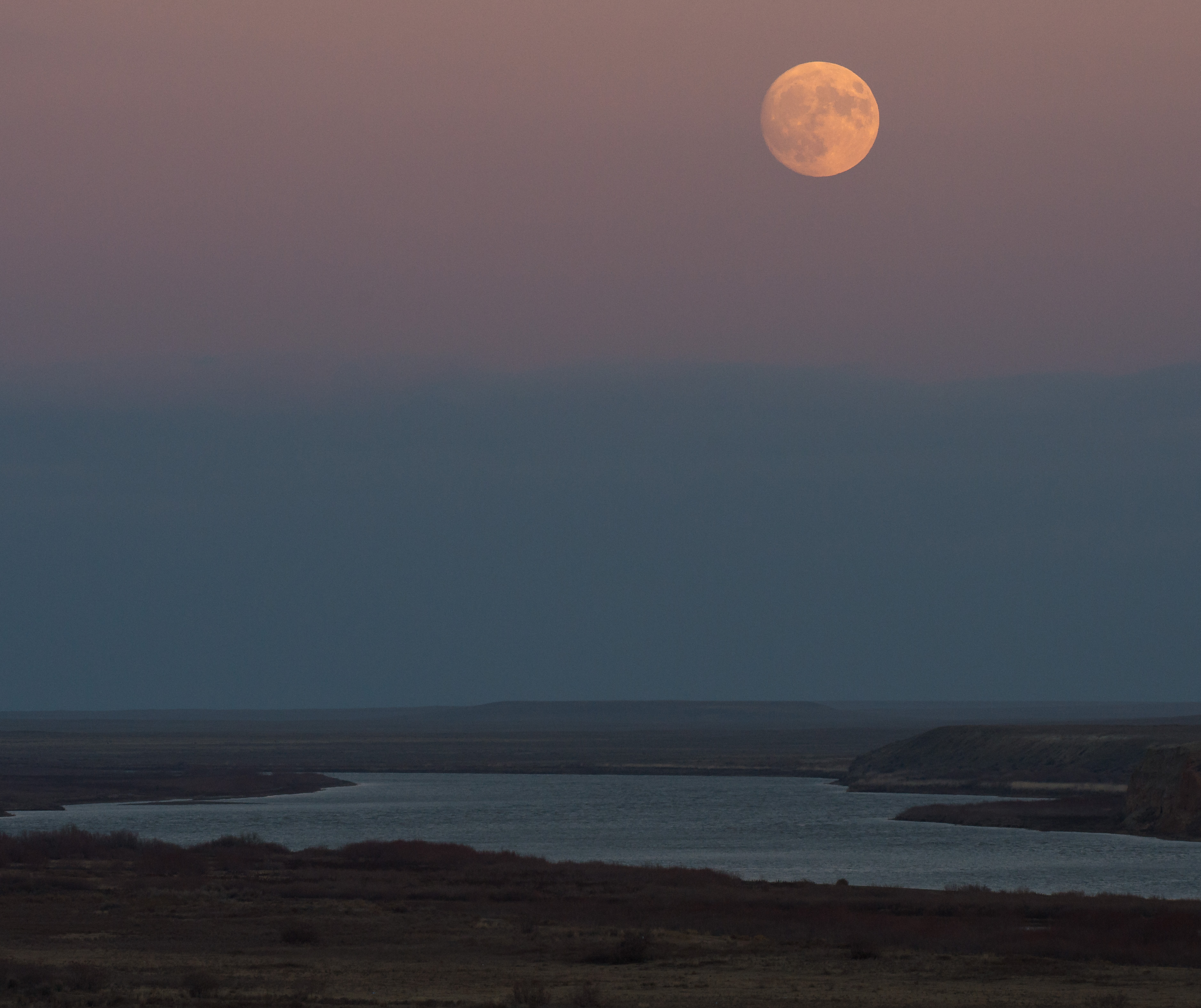 pink Moon in a pink sky, over a hazy blue river. 
