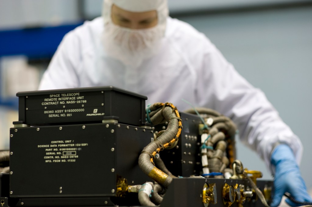 Man in a white clean room suit looking at Hubble instruments in black boxes with wires coming out of the sides.