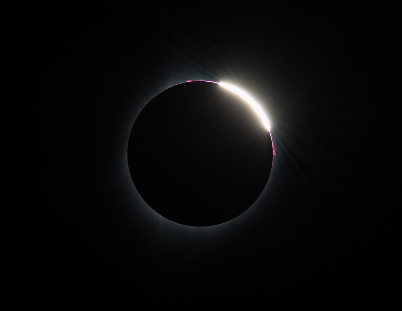 The eclipsed Sun, appearing as a black circle with a faint white glow. On the upper area, bright white and pink areas peak out from behind the Moon.