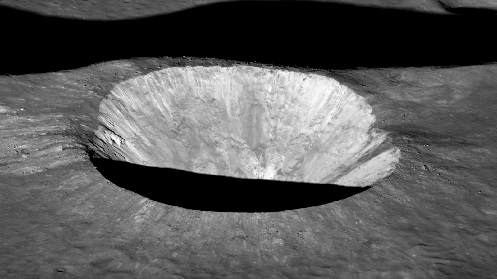 Dramatic close-up of a brightly lit lunar crater, seen at oblique angle. The crater's edge casts a deep shadow onto a thin slice of its floor.
