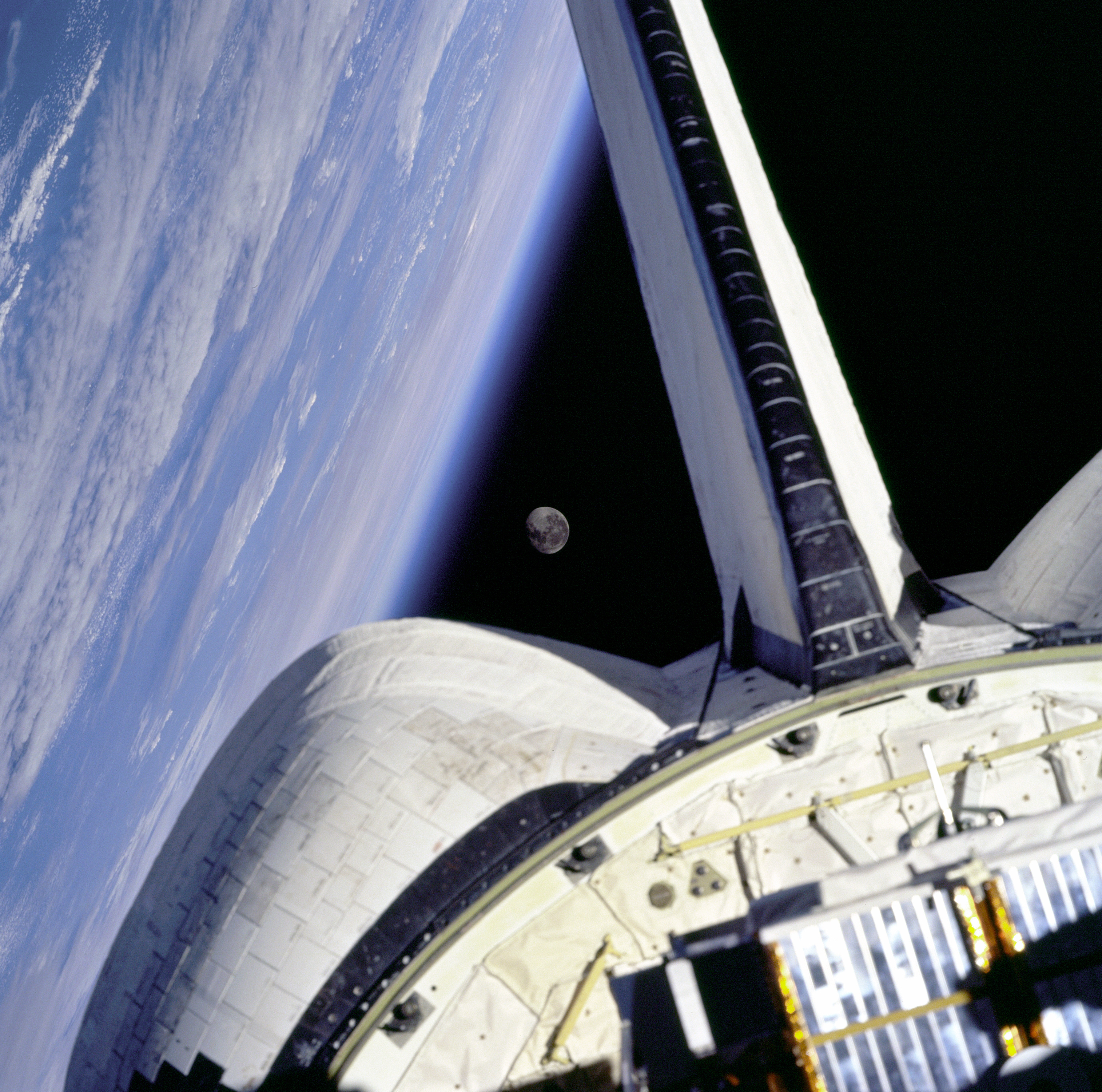 Earth's horizon looms on the left in the background. In the foreground, a spacecraft's tail parts fill the bottom of the image. Between the two is a triangle of black space containing the Moon. 
