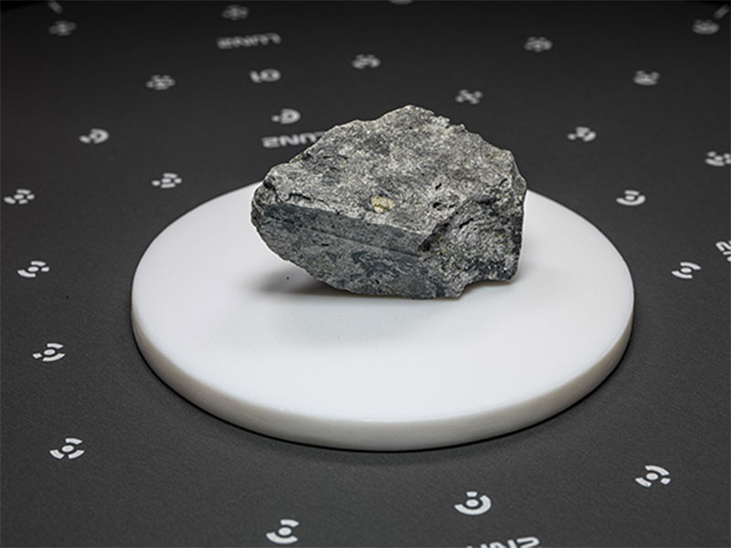 Black background, dotted with white spots and a circular white tray in the center with a piece of space rock sitting atop.