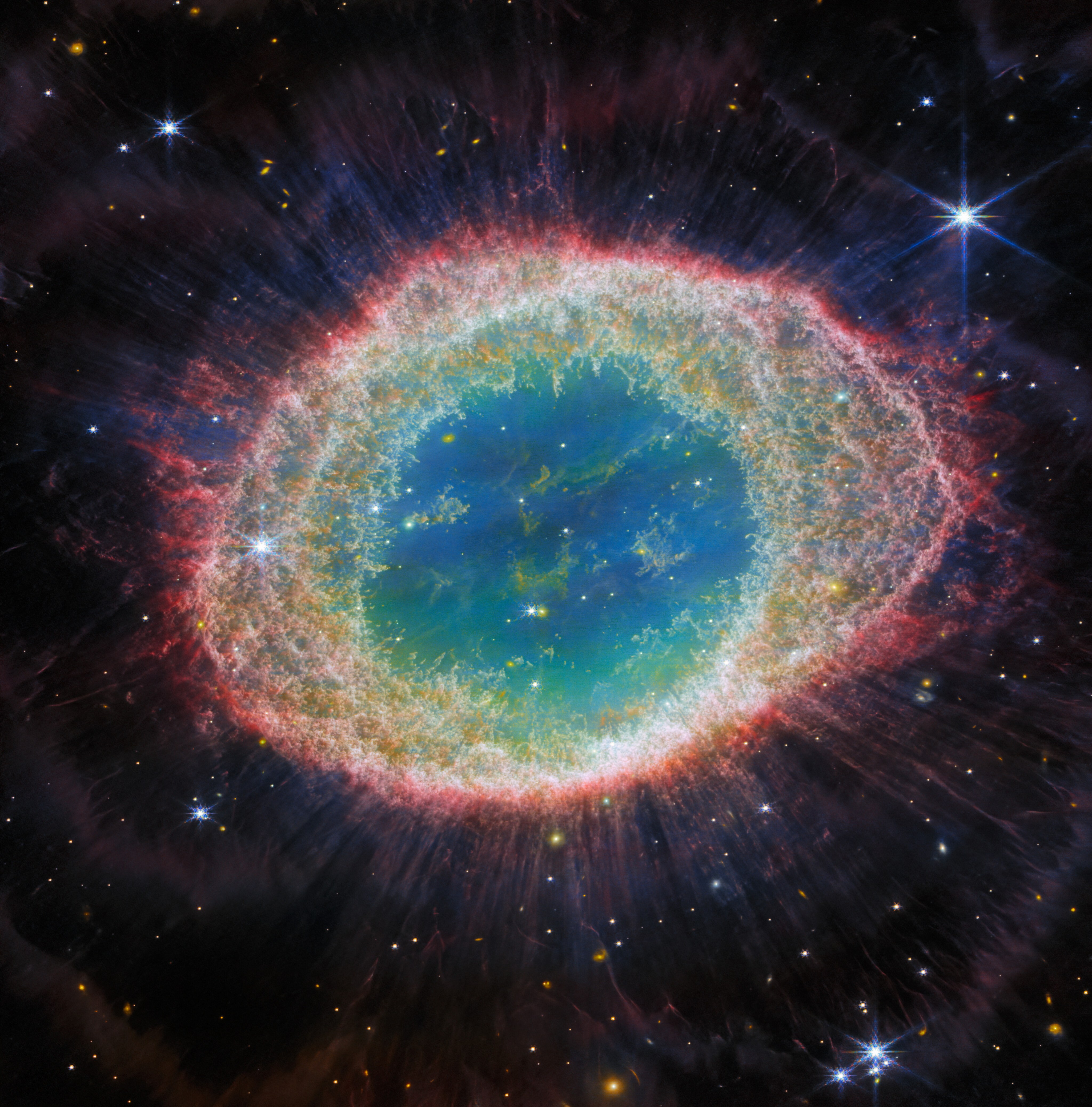 NIRCAM IMAGE: This image of the Ring Nebula appears as a distorted doughnut. The nebula’s inner cavity hosts shades of blue and green, while the detailed ring transitions through shades of orange in the inner regions and pink in the outer region. The ring’s inner region has distinct filament elements.