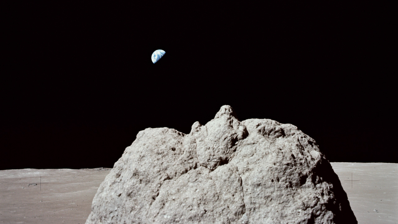 Moon terrain with a large boulder poking up from below the field of view in the foreground and Earth overhead in the background