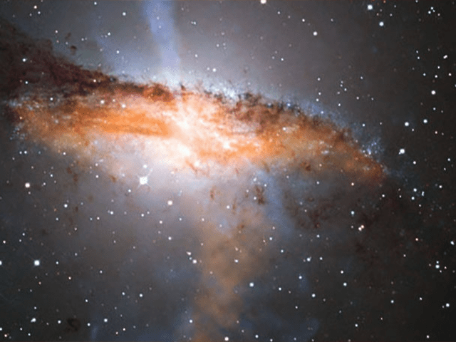 Image of a brightly, colorful cloud of material–-a galaxy–in dark, starry space. The galaxy is lit from within, giving it a white and orange glow with dark purple shadows. Hazy streaks of light emerge from its top and bottom.