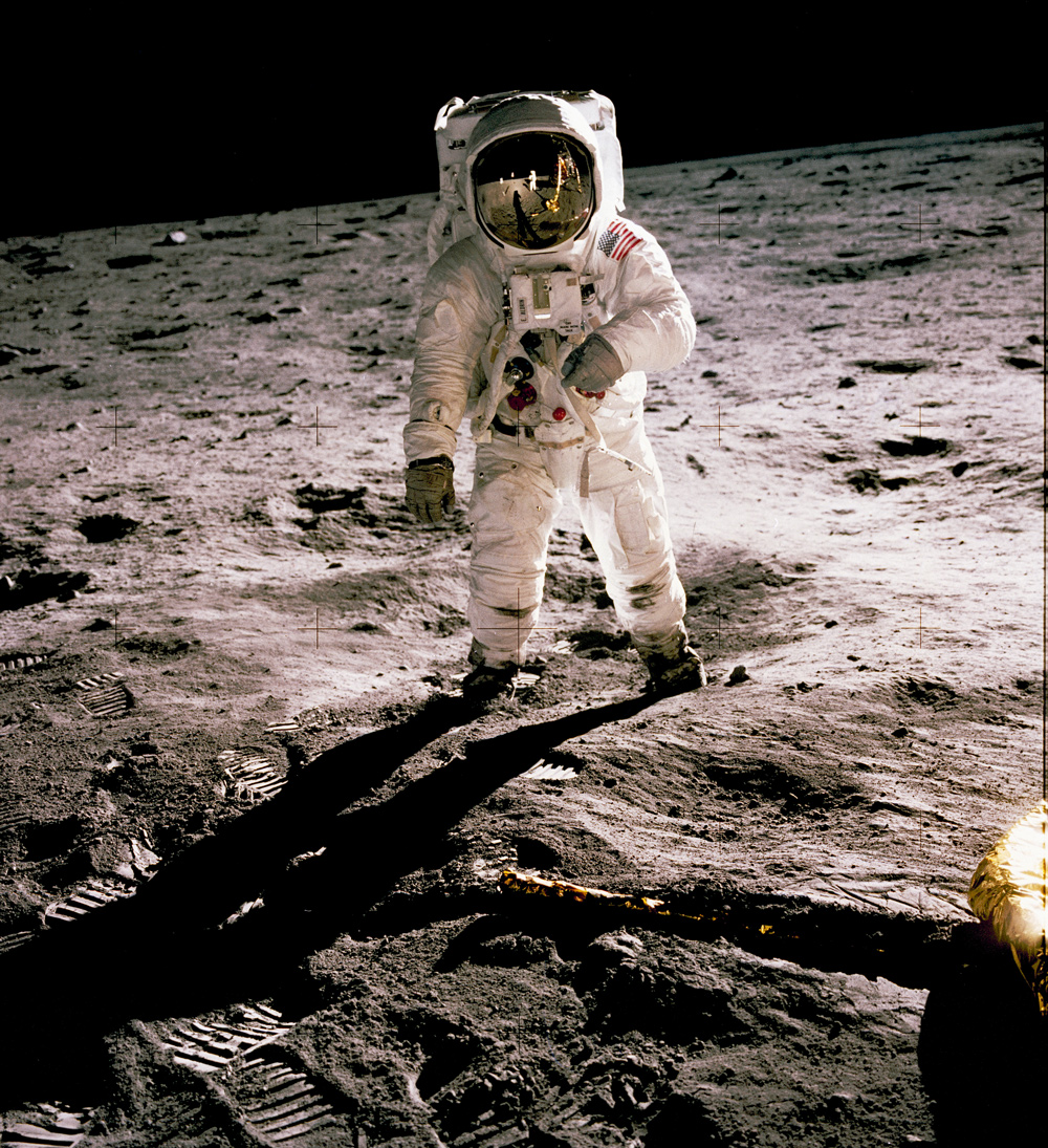 Astronaut on the Moon, facing the viewer. Another astronaut and a spacecraft are reflected in his helmet visor.