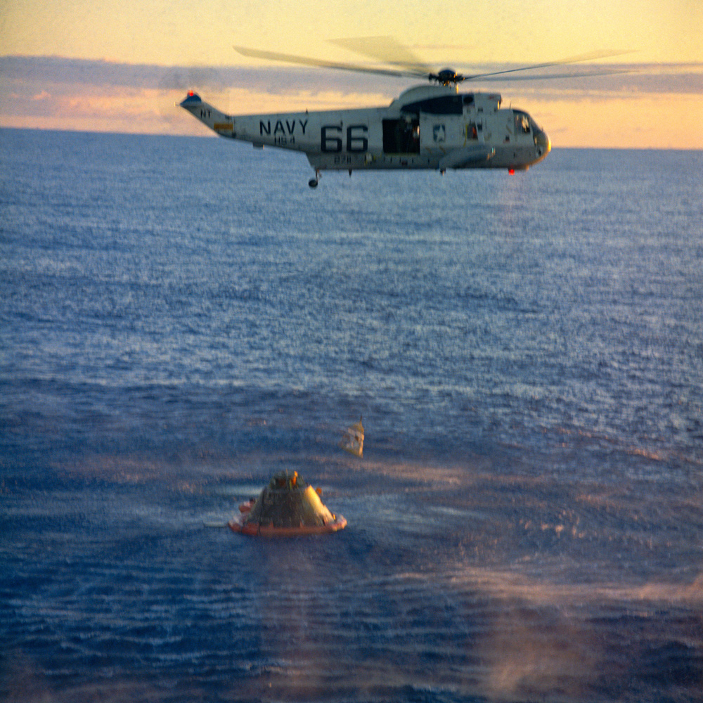 Helicopter lifting astronaut from recovery capsule over Pacific ocean, under a yellow-orange sky