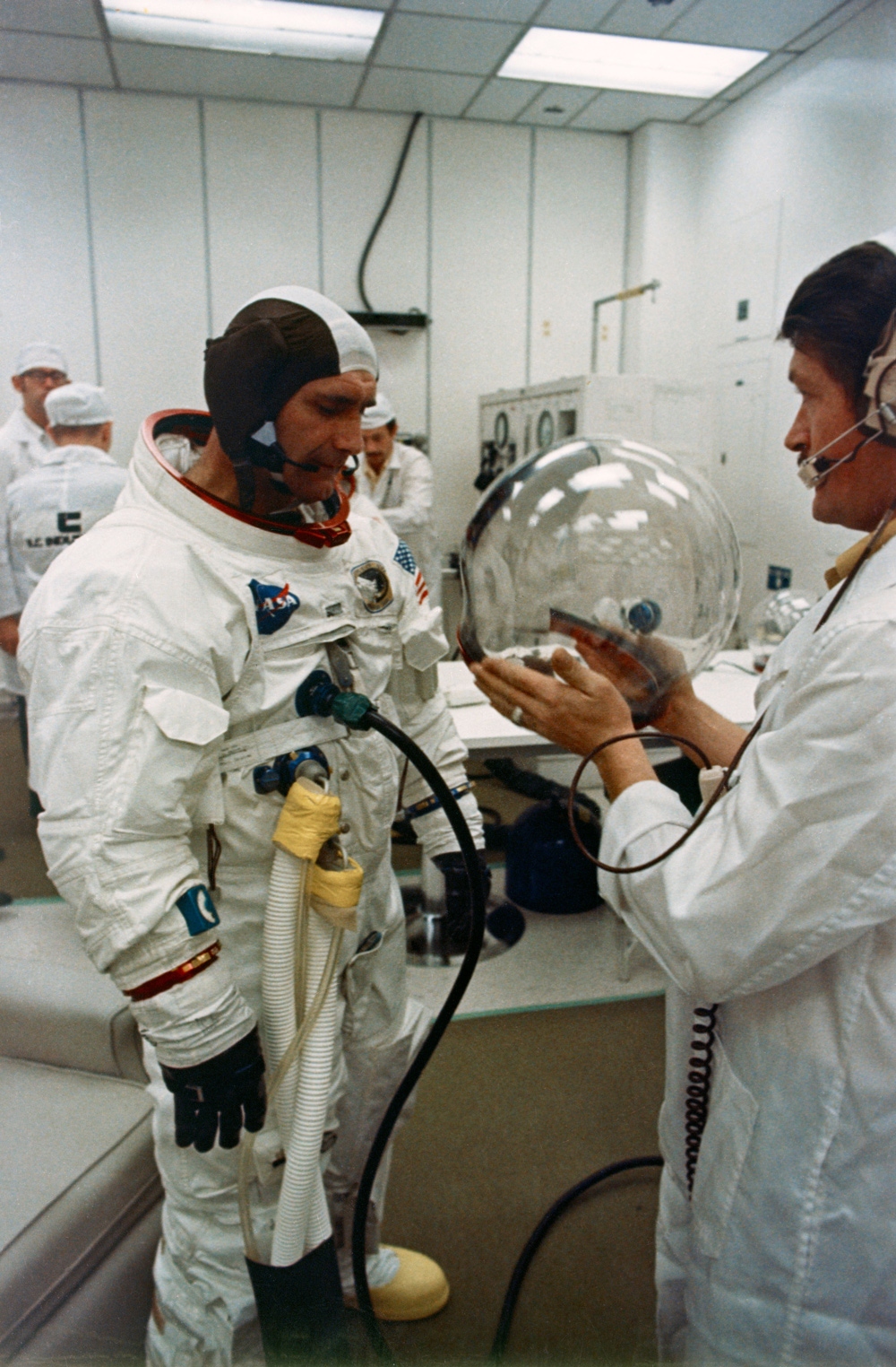 Astronaut preparing to don helmet with help of another person, in a white-walled room