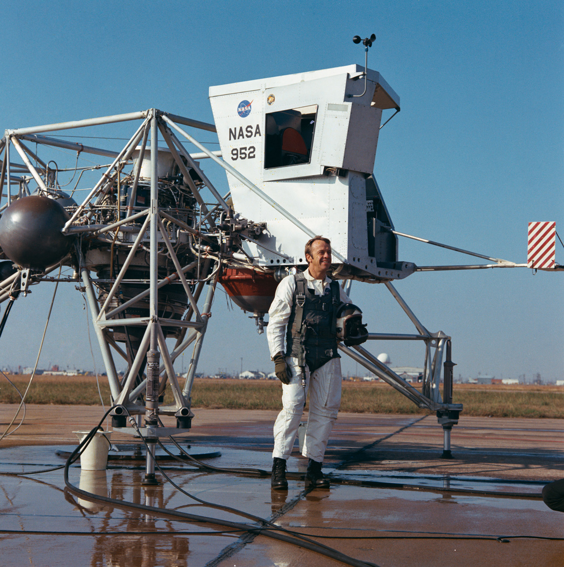 Astronaut standing next to training vehicle, smiling, on wet pavement, under a blue sky.