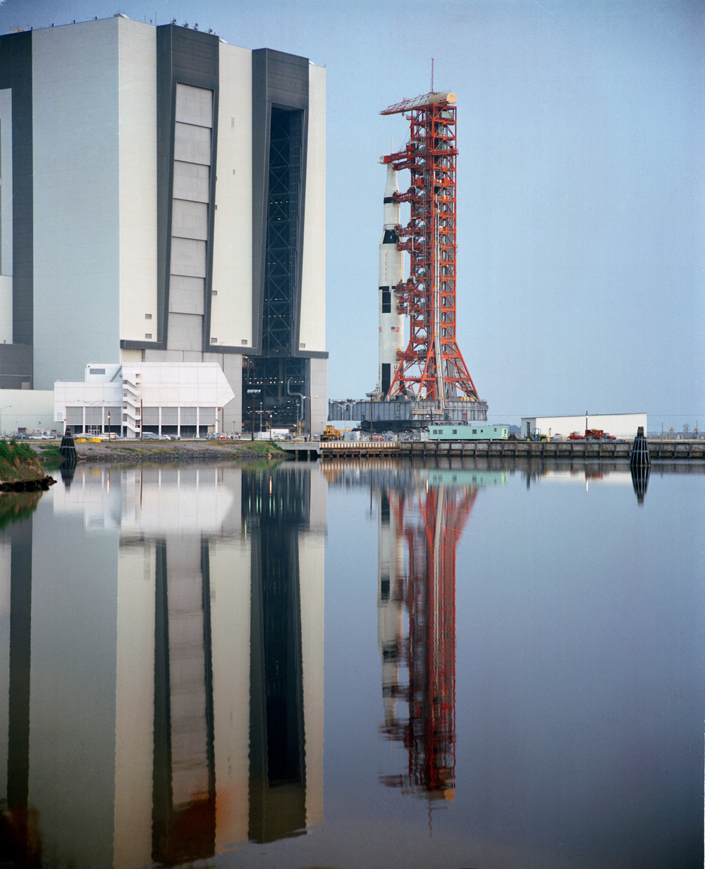 View of Apollo 15 space vehicle, and tall adjacent building, reflected in a smooth body of water.