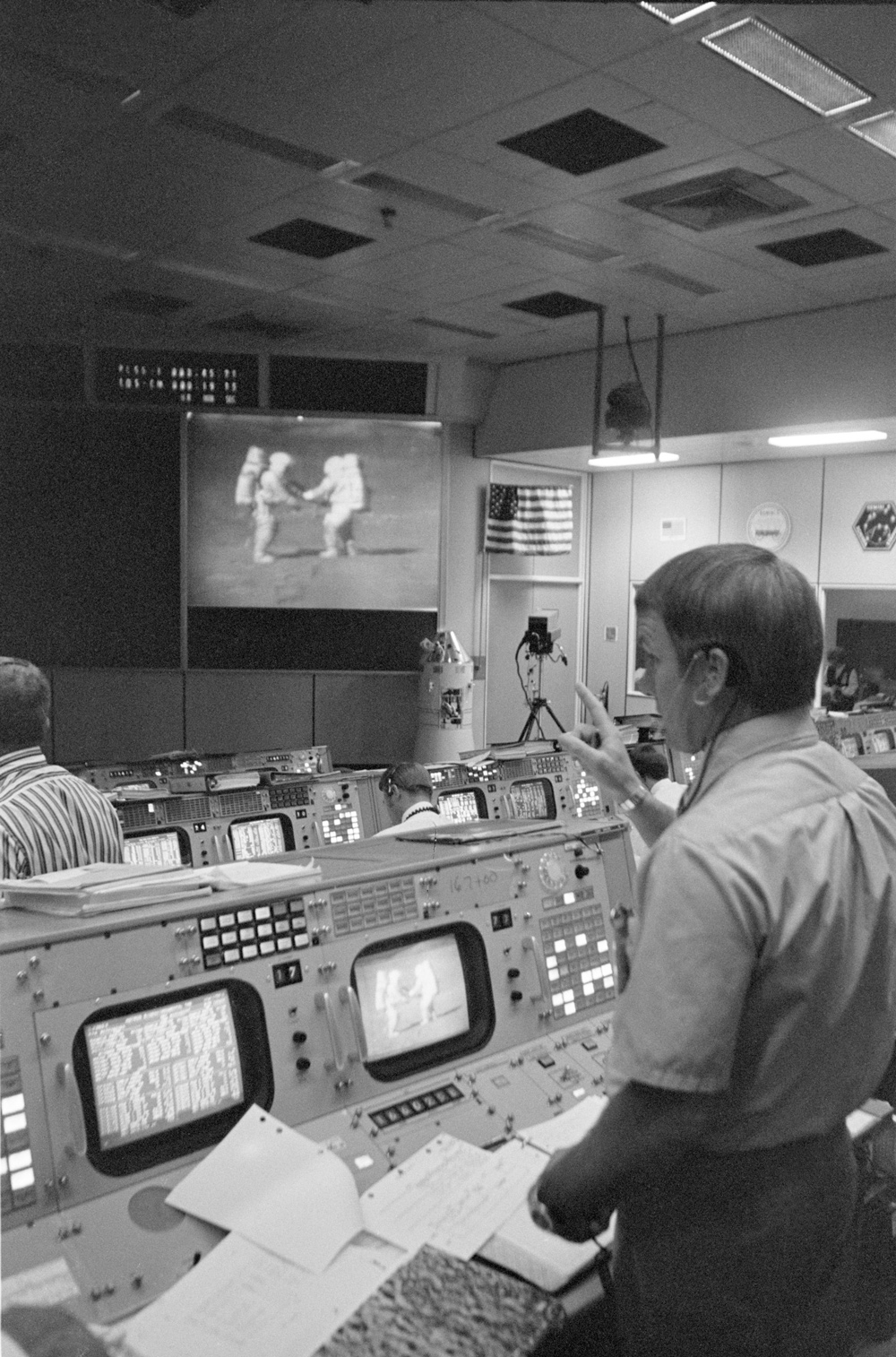 Apollo 15 flight director in Mission Control Center, surrounded by old-fashioned computer screens and interfaces. He raises a hand to point towards an image of astronauts on the Moon projected on the wall.