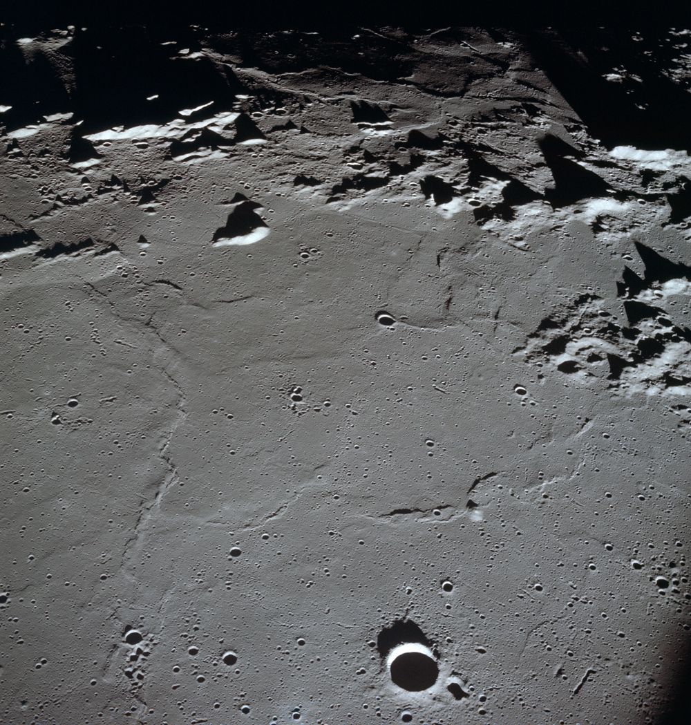 Moon's surface. At the bottom of the image, a sharply defined, round crater with darkly shadowed floor. At the top edge, mountain ridges cast deep shadows away from the camera.  