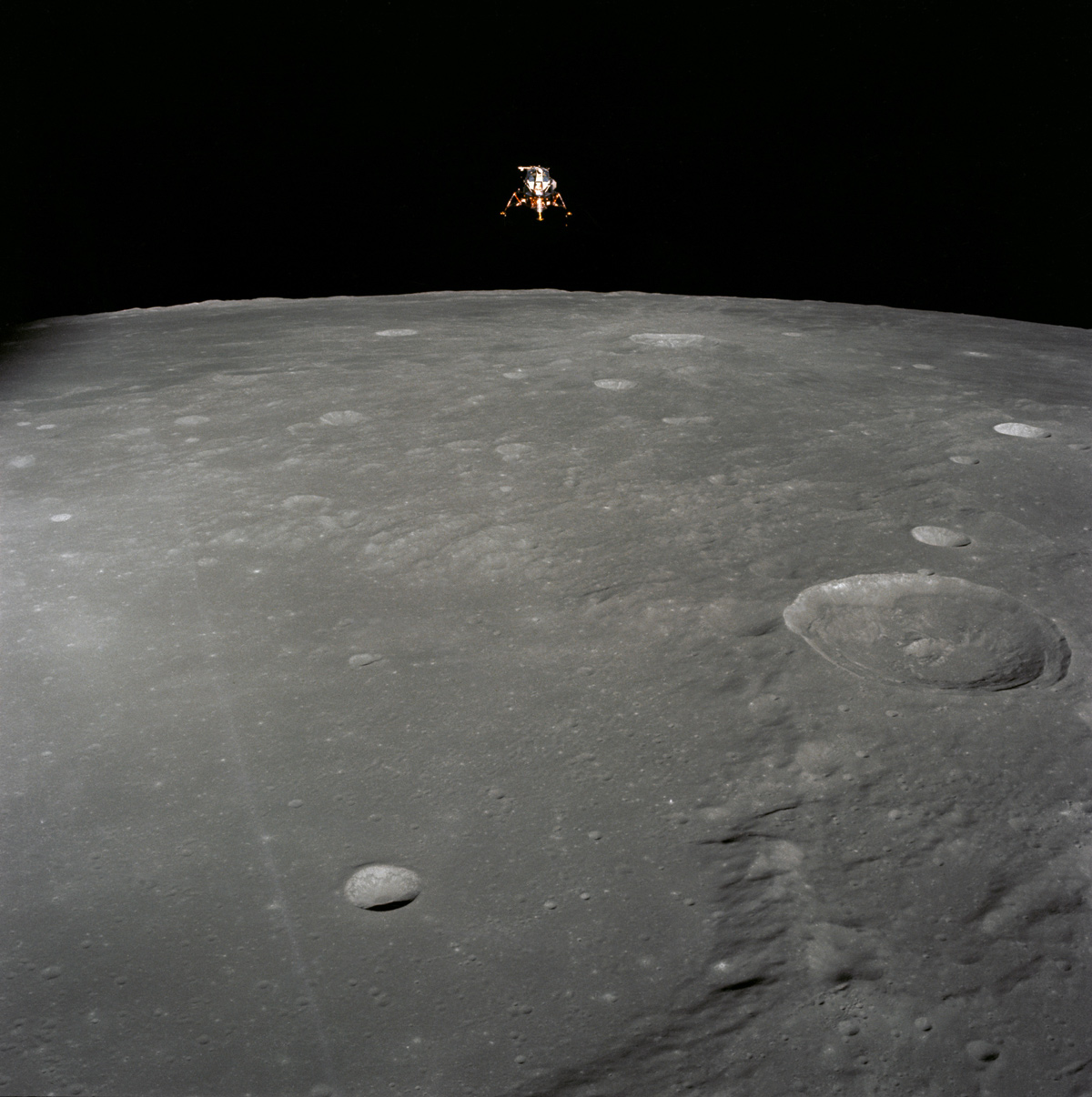 The Apollo 12 Lunar Module in lunar orbit, visible in space over the Moon's horizon. On the lunar surface below, a row of craters and hills extending towards the viewer.
