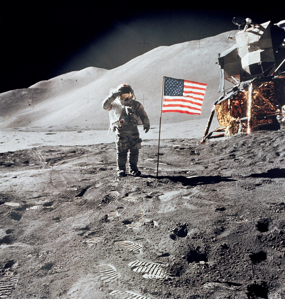 Astronaut giving salute beside American flag on the Moon, with lander in the background. In the foreground: bootprints in fine, grey dust.