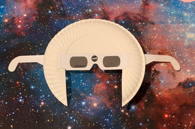 white solar eclipse viewing glasses with a black nasa insignia in the middle attached to a paper plate and red and blue galaxy background