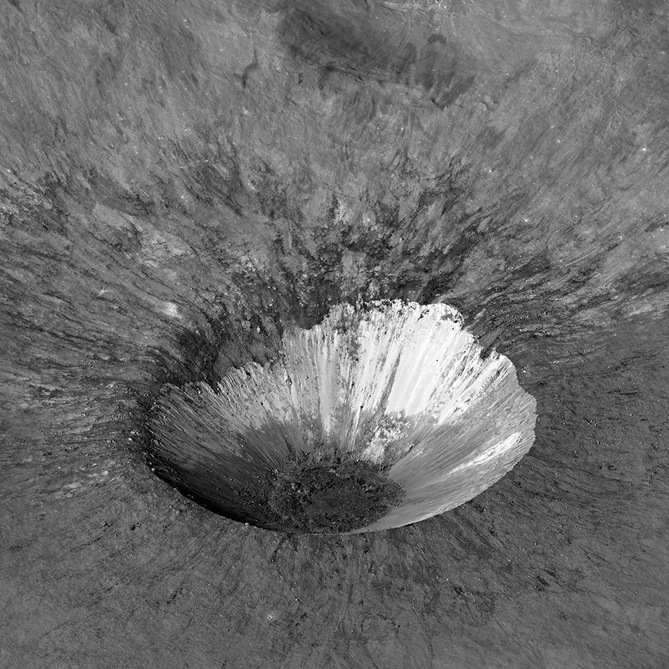Sharply defined crater with rugged, asymmetrical edge and bright areas visible where surface material has been excavated on impact