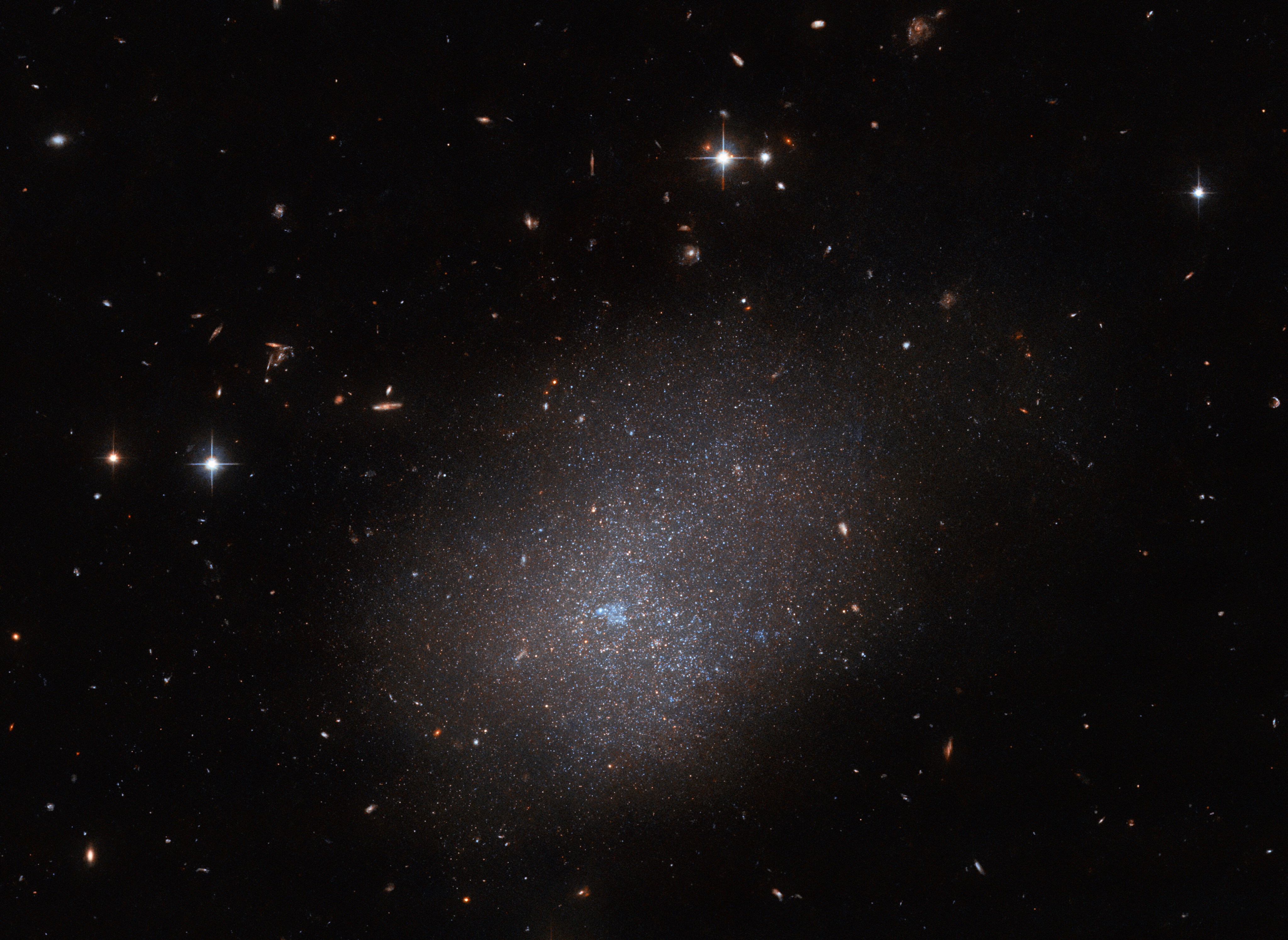 An irregular galaxy that resembles the shape of a cloud. It is made of many tiny stars all clumped together, surrounded in a diffuse light. In the central, brightest part there is a bubble of blue gas. The galaxy is surrounded by mostly very small and faint objects, though there are bright stars above and to the left of it, and a string of galaxies nearby.