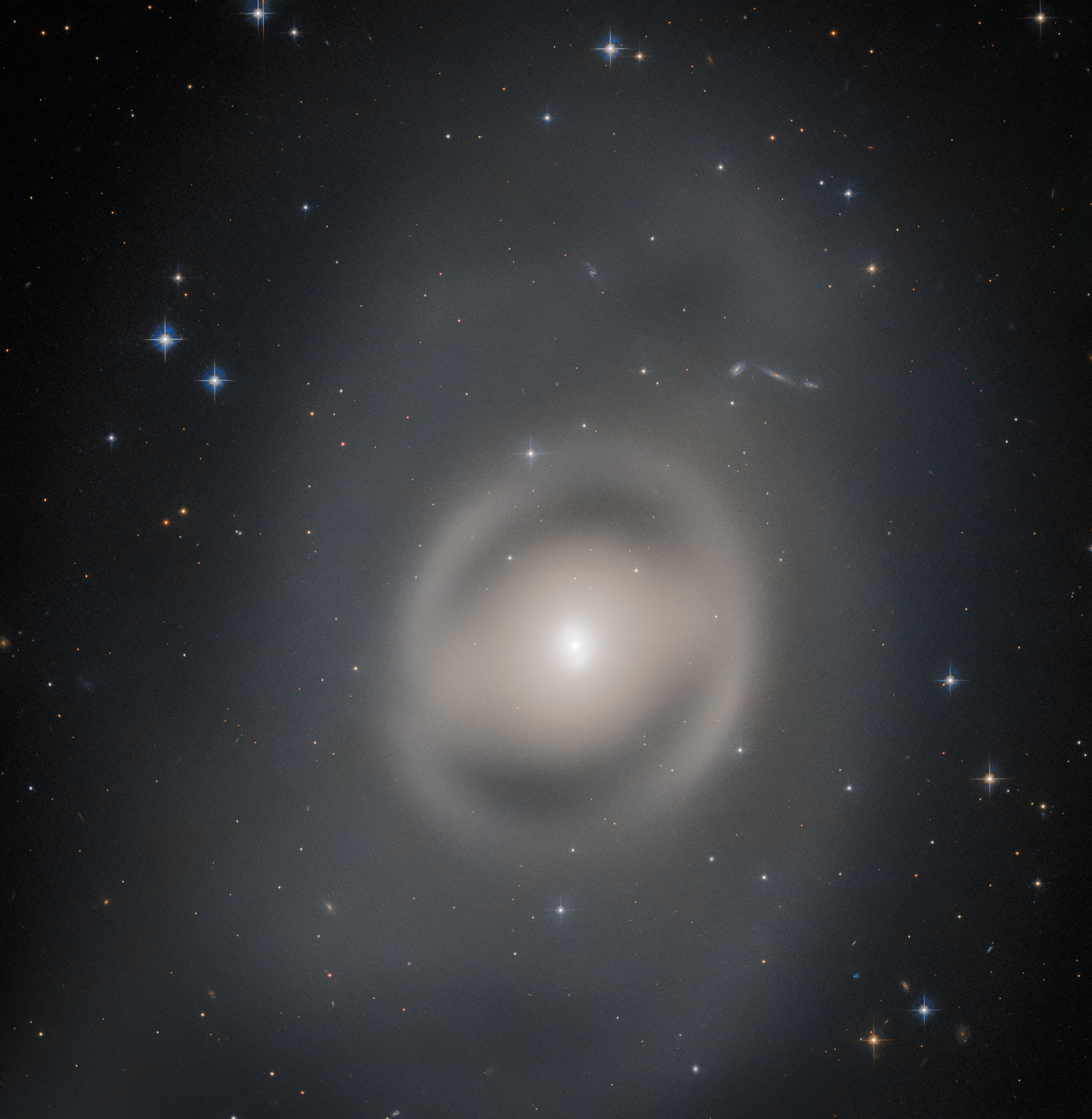 A galaxy, large and occupying most of the view from the center. The whole galaxy is made of smooth, diffuse light. The galaxy is surrounded by a smoky gray halo. Many stars shine around the galaxy, on a black background.